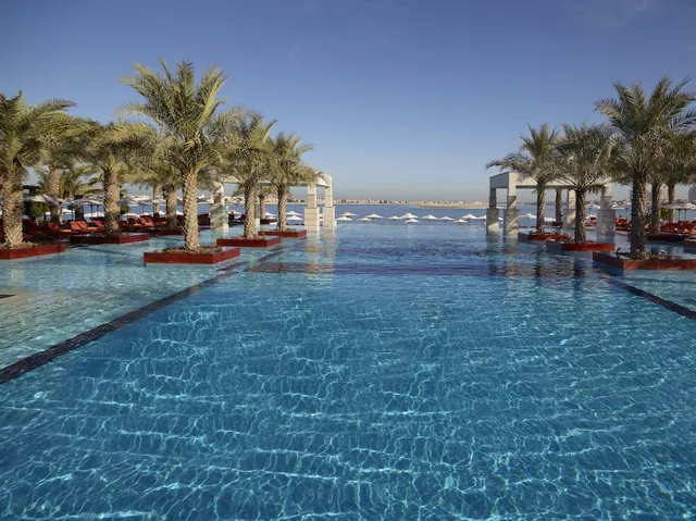 An infinity-edge swimming pool lined with palm trees and sun loungers, with the sea and white umbrellas visible beyond the far edge.