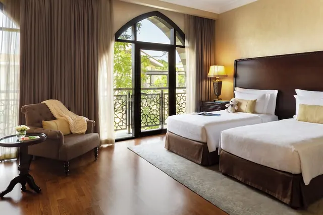A bedroom with two single beds, an armchair and side table, and an arched glass door opening onto a balcony with trees beyond.