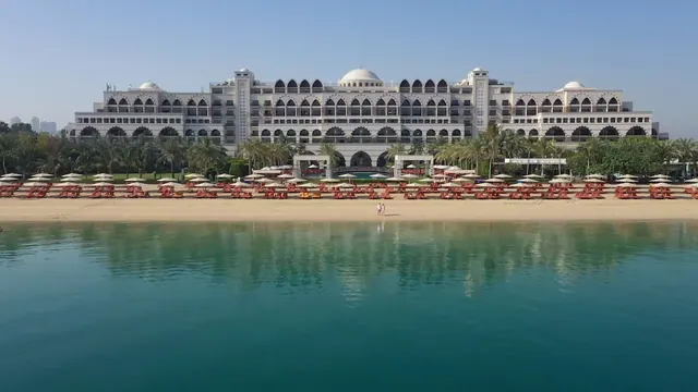 Aerial view of a large white hotel with domes and arched windows behind a sandy beach lined with red loungers and umbrellas, with turquoise sea in the foreground.