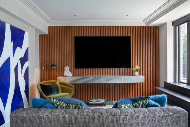 A living area with a wall-mounted television on a slatted wood wall, a marble console, a grey tufted sofa and a yellow velvet armchair.
