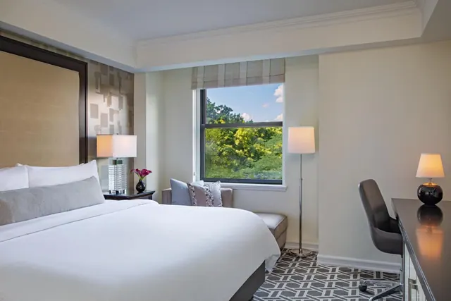 A hotel bedroom with a made king bed, a seating area by a window looking out onto trees, and a desk with a chair on the right.