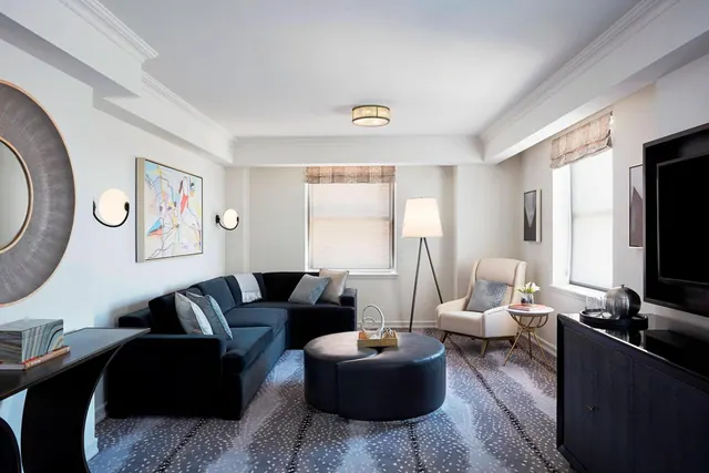 A living area with a navy sectional sofa, a round leather ottoman, an armchair, a floor lamp and a wall-mounted television.