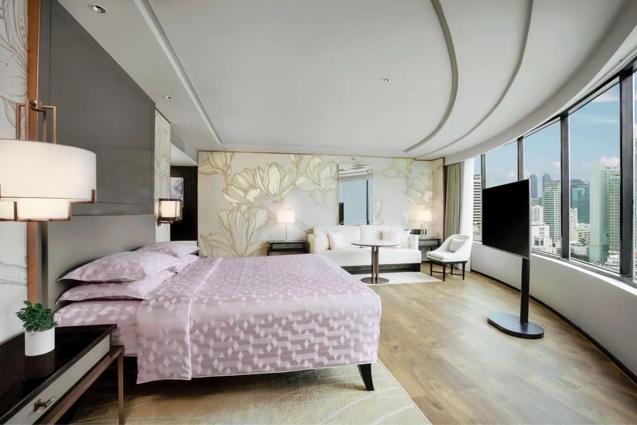 A bedroom with a made king bed, a white sofa and round table, and a freestanding television beside curved floor-to-ceiling windows overlooking city high-rise buildings.