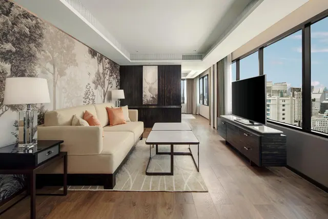 A living room with a beige sofa, a tree mural on the wall, a low coffee table, and a wall-mounted television beside floor-to-ceiling windows that look out over a city skyline.