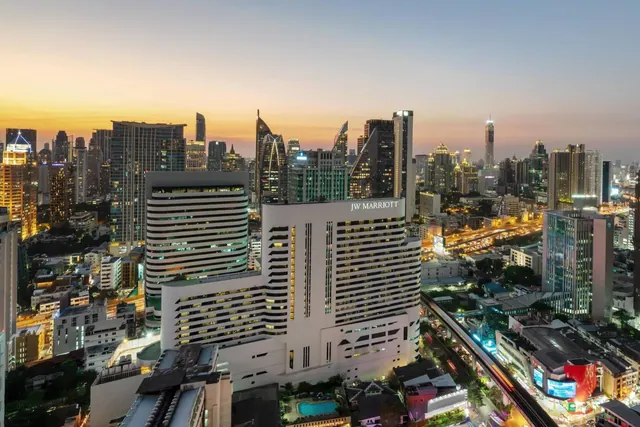 Aerial dusk view over Bangkok showing the JW Marriott hotel tower, surrounding skyscrapers, a small pool and an elevated rail line.