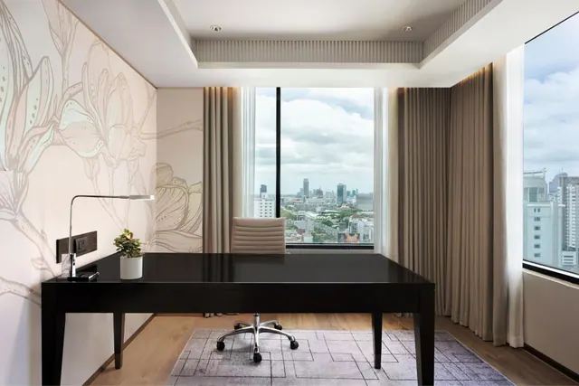 A black desk and chair stand in a room with floor-to-ceiling windows overlooking a city skyline.