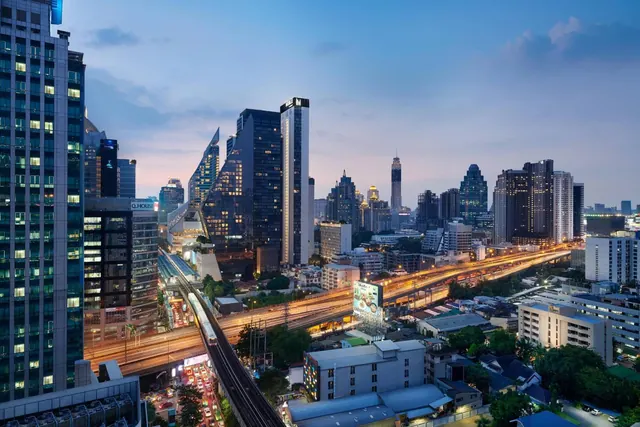 Dusk view over Bangkok's skyline showing illuminated high-rise towers, an elevated expressway with light trails, and a Skytrain line in the foreground.