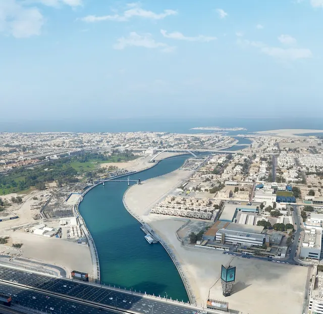 Aerial view of the Dubai Water Canal curving past low-rise residential and commercial buildings toward the sea on the horizon.