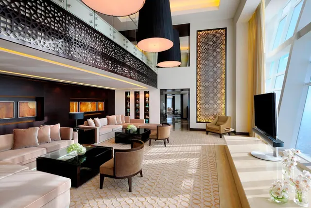 A double-height living area with cream sofas, armchairs, dark coffee tables, pendant lamps, and a television on a stand beside tall windows.