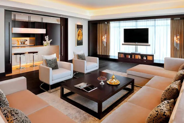 A staged living area in a hotel suite with a sofa set, armchairs, a coffee table, a wall-mounted television and a kitchen with bar stools in the background.