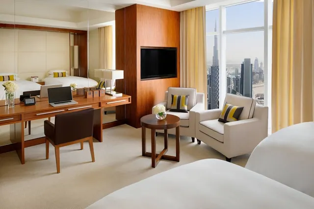 A hotel bedroom with a bed, a wooden desk, two armchairs and a wall-mounted television, with floor-to-ceiling windows showing the Burj Khalifa and the city skyline.