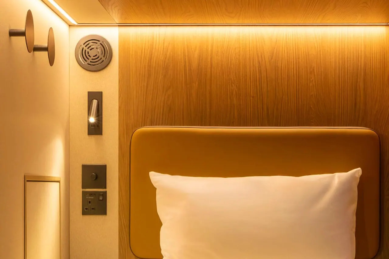 A made bed with a white pillow and mustard upholstered headboard sits against warm wood panelling, beside a reading light, wall sockets and a ceiling speaker grille.