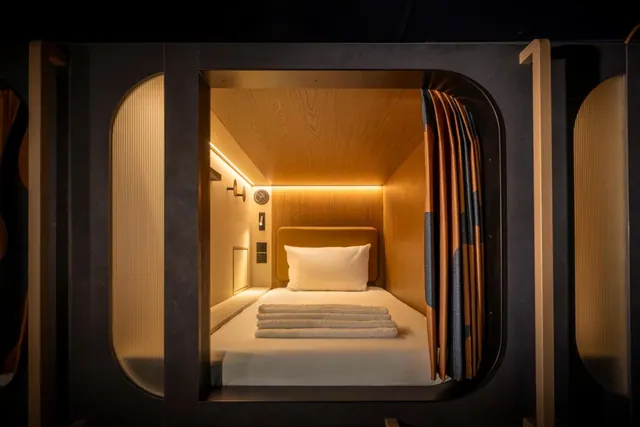 A single bed with a pillow and folded towels sits inside a warmly lit wood-panelled sleeping pod with a patterned curtain.
