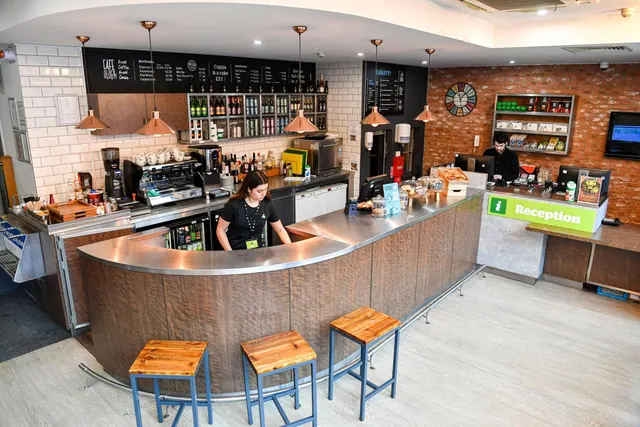 A café counter with wooden stools and a staff member behind it sits beside a reception desk with a green i Reception sign.