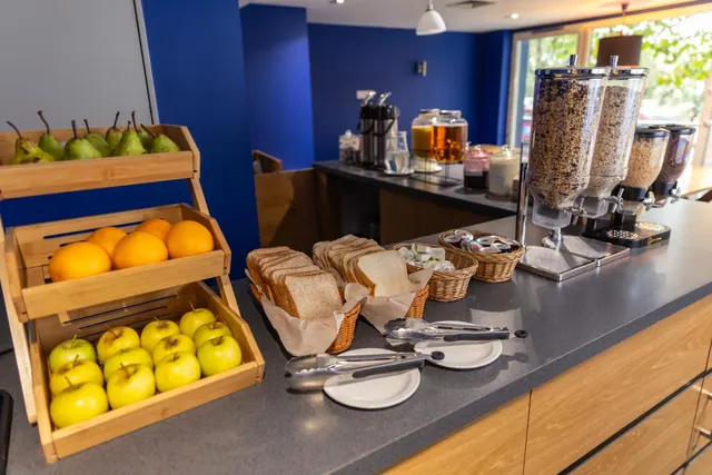 A breakfast buffet counter with baskets of pears, oranges and apples, sliced bread, cereal dispensers, and stacked plates and tongs.