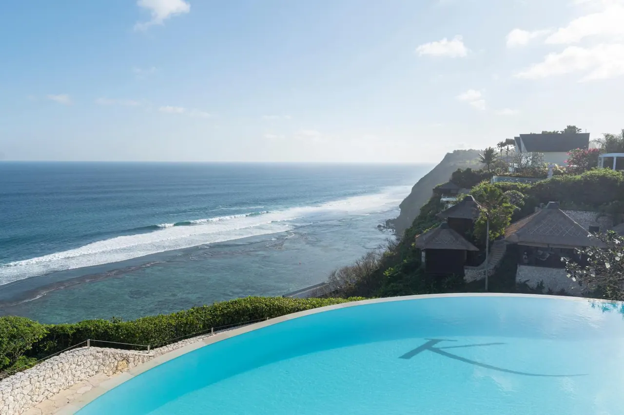 An infinity-edge pool overlooks the ocean, with waves breaking along the coastline and cliffside villas to the right.