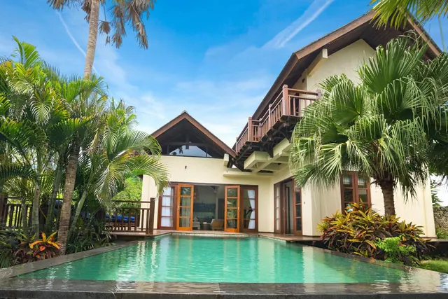 Exterior of a two-storey villa with an upper balcony, a swimming pool in front and palm trees around it.