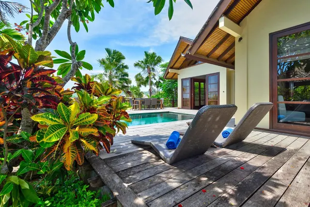 A timber deck with two wicker sun loungers and folded blue towels sits beside a small swimming pool and a villa with a timber-ceilinged veranda.
