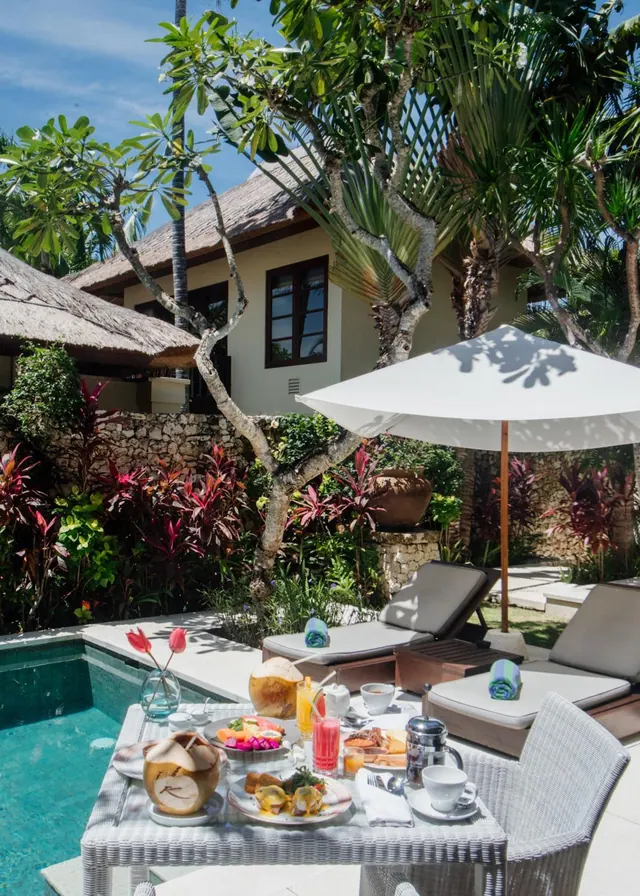 A breakfast table with plates, drinks and a coconut sits beside a small private pool with sun loungers and a white parasol.