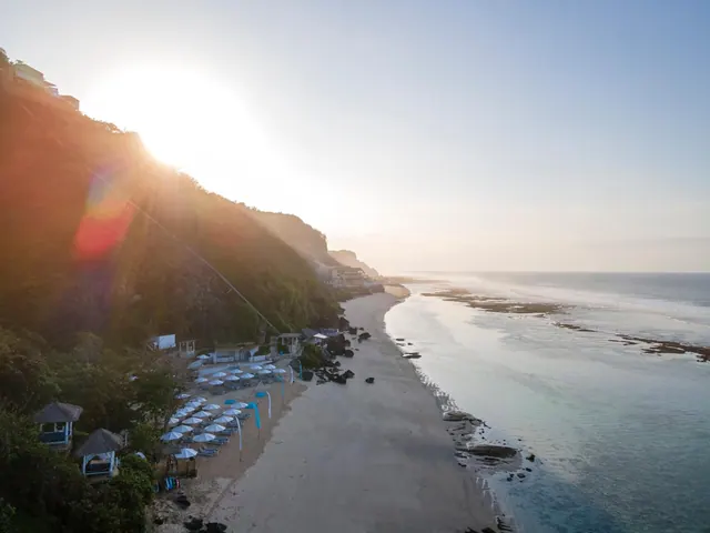 Aerial view of a sandy beach with white parasol umbrellas and sun loungers at the foot of a cliff, with rocks and the sea visible under low sun.