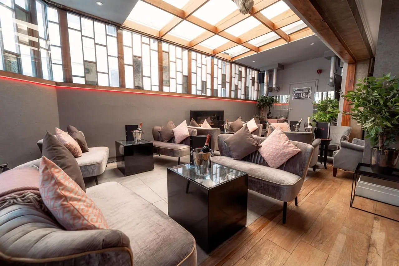 A lounge with grey velvet sofas, pink cushions, black side tables and a champagne bucket, under a glazed skylight roof.