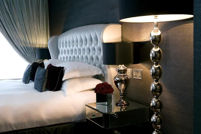 A bedroom with a made king bed, a tufted grey headboard, dark cushions, a mirrored nightstand holding a red rose arrangement, and a tall chrome lamp.