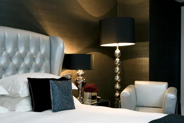 A bedroom with white linen on the bed, a tufted silver leather headboard, a silver leather armchair, and floor and table lamps against dark textured wallpaper.
