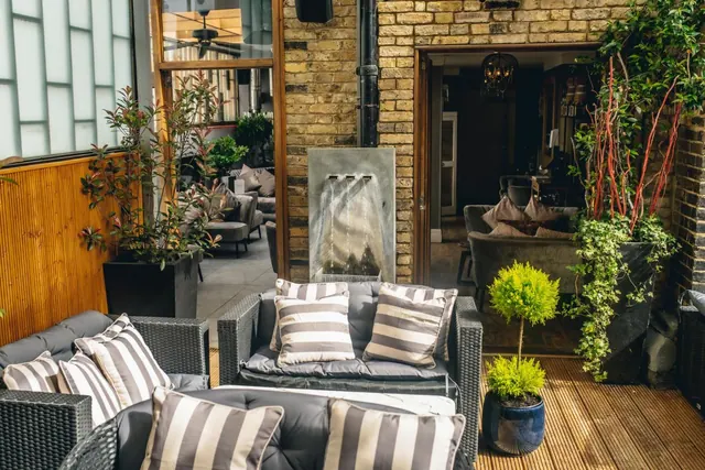 An outdoor terrace with grey wicker sofas and striped cushions on wooden decking, beside a brick wall with a water feature and potted plants.