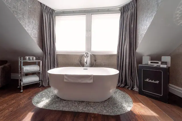 A freestanding white bathtub stands on a grey stone-effect rug in front of a bright window with grey curtains, beside a towel trolley and a Marshall amplifier cabinet.