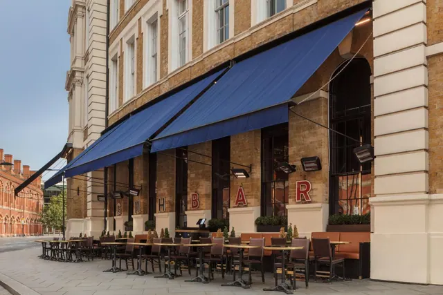 Exterior of a brick building with blue awnings over an outdoor seating area of tables and chairs on the pavement, with red lettering on the facade.