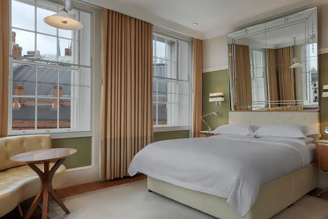 A bedroom with a made king bed, a tufted leather banquette beside a round wooden table, and tall sash windows looking out over rooftops and chimneys.