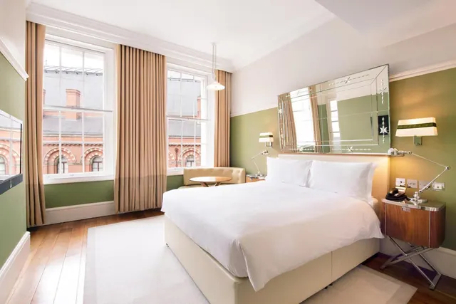 A bedroom with a large white bed, green walls, a small sofa and round table by the windows, and red brick buildings visible outside.