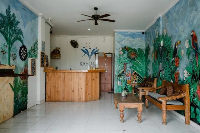 A lobby with a wooden reception counter, jungle-themed wall murals, wooden benches, a low wooden coffee table and a ceiling fan above a tiled floor.