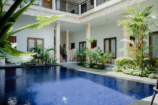 A rectangular blue swimming pool sits in a courtyard surrounded by two-storey white buildings, palm trees and landscaped planting.