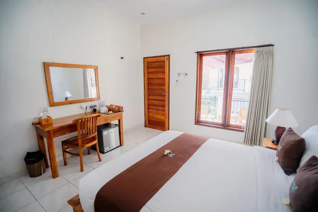 A bedroom with a double bed, a wooden desk with a mirror and chair, a small fridge, and a window looking onto a neighbouring balcony.