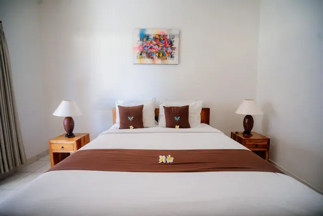 A made double bed with white linens, a brown runner decorated with frangipani flowers and brown cushions, flanked by two wooden nightstands with lamps and an abstract painting on the wall.