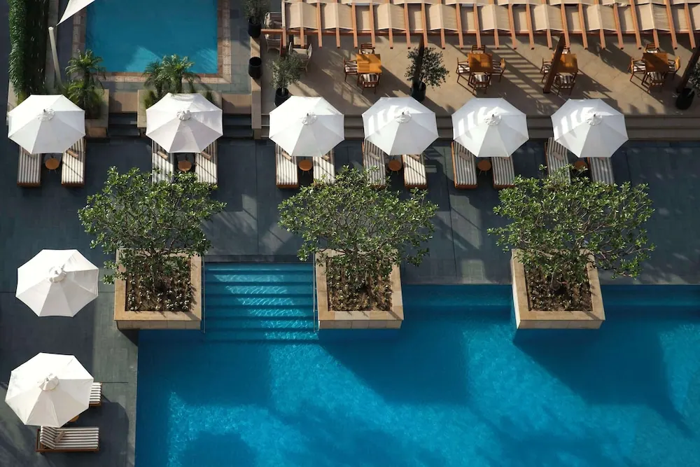 Aerial view of a large swimming pool with white umbrellas, sun loungers, potted trees and outdoor dining tables under a wooden pergola.