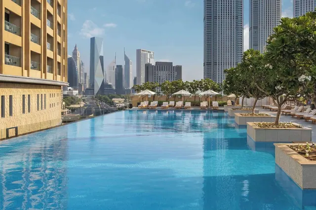 A long outdoor swimming pool with sun loungers and white umbrellas beside it, with a city skyline of high-rise towers in the background.