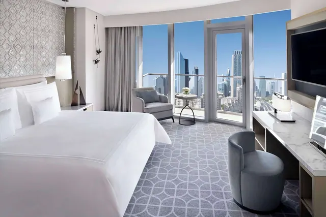 A hotel bedroom with a king bed, armchair, desk and wall-mounted television, with floor-to-ceiling windows and a balcony door looking out over city towers.