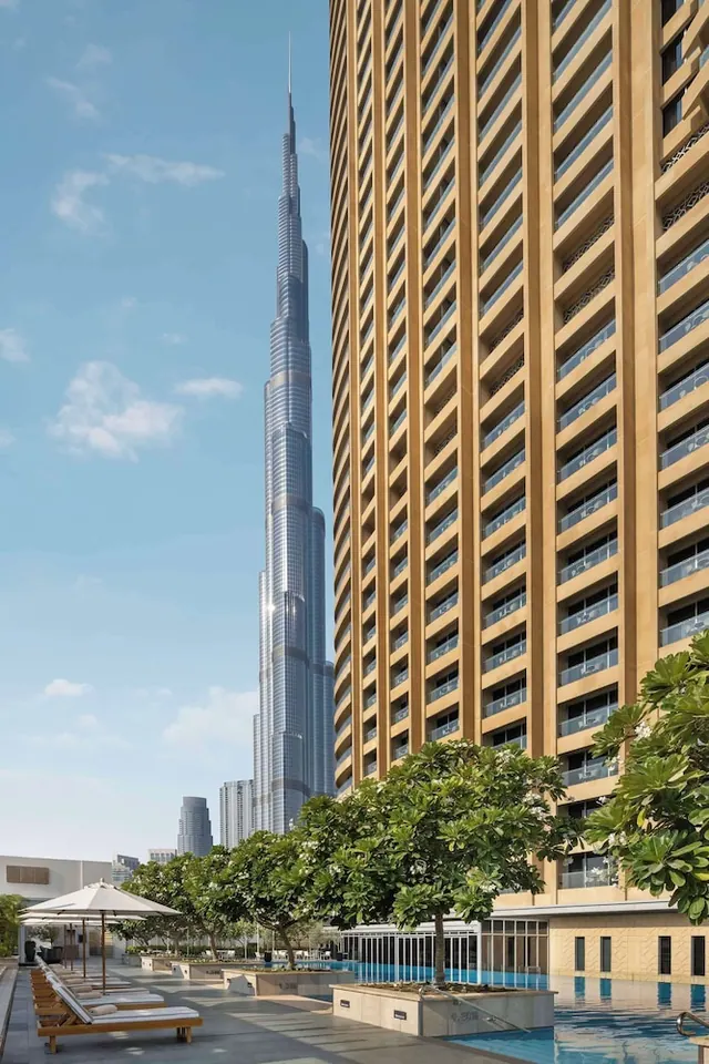 The Burj Khalifa rises behind a tan high-rise hotel tower, with sun loungers and umbrellas on the pool deck in the foreground.