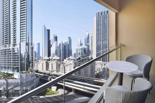 A small round marble table and two wicker chairs sit on a balcony with a glass railing, overlooking high-rise towers and a road below.
