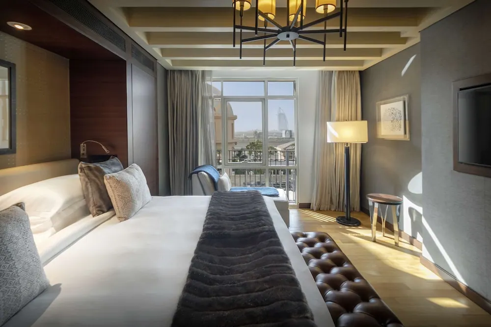 A king bed with a fur throw and a tufted leather bench at its foot faces a balcony door with a view of Burj Al Arab.