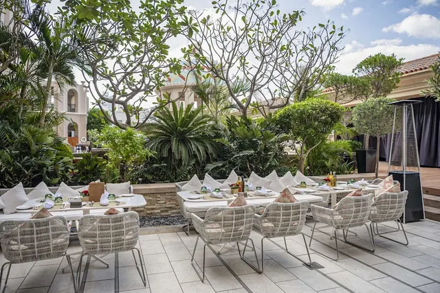 An outdoor terrace dining area with white wicker chairs and set tables, surrounded by palm trees and tropical garden plants.