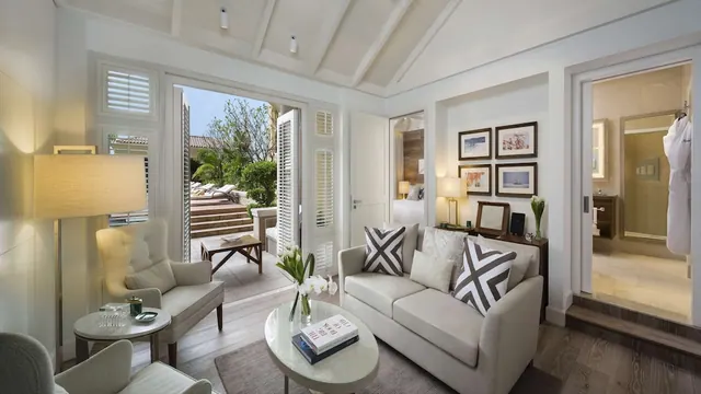 A bright living area with a sofa, armchairs and a round coffee table, with open doors onto an outdoor terrace with loungers and greenery, and a bedroom and bathroom visible beyond.