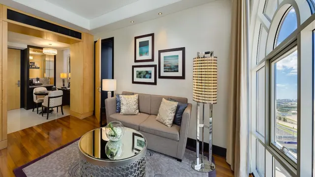 A hotel suite living area with a grey sofa, a floor lamp, framed wall prints and a large arched window looking out over distant buildings and greenery.