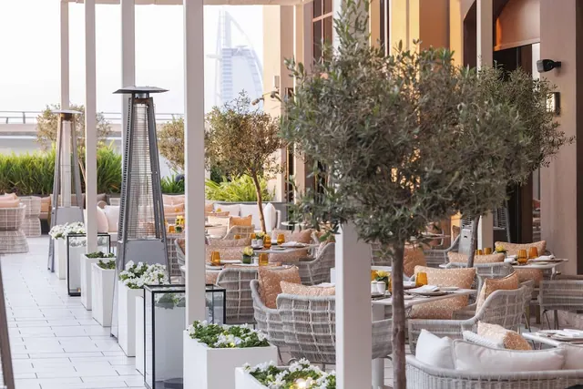 An outdoor dining terrace with olive trees, wicker armchairs at set tables, patio heaters and white planters, with the Burj Al Arab visible in the background.