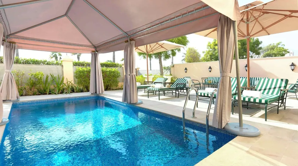 A rectangular swimming pool with blue water sits beside a canopy, striped sun loungers and parasol umbrellas on a tiled deck.