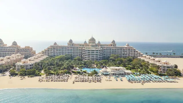 Aerial view of a large white resort with a central pool, rows of beach umbrellas and loungers on the sand, and the sea in the foreground and background.