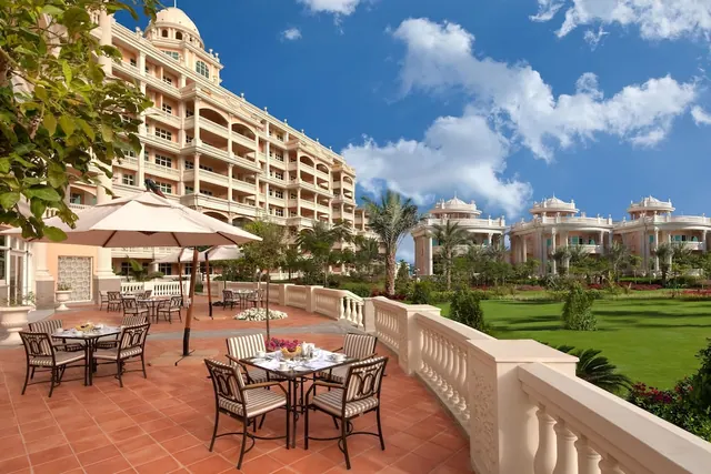A terrace with set outdoor dining tables, parasols and chairs overlooks a lawn, palm trees and pale pink buildings under a blue sky.