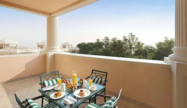 A breakfast table with fruit, orange juice, pancakes and a croissant sits on a covered balcony with green striped chairs, overlooking trees and rooftops.